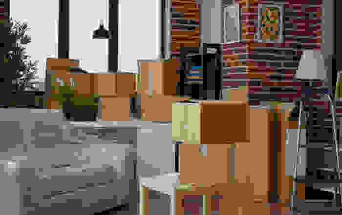 Living room with moving boxes and furniture wrapped in plastic, brick wall background.