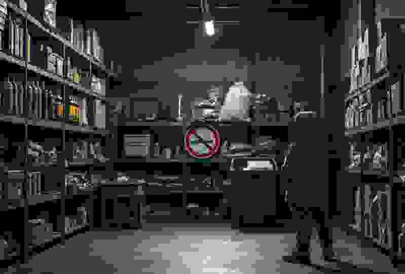 A child in a hooded jacket stands in a cluttered storage room, surrounded by shelves of supplies and a no-weapon sign.