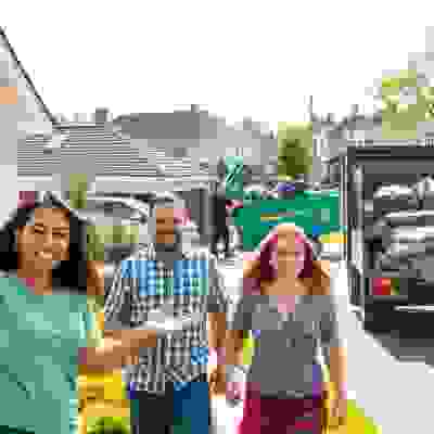 A group of people stands outside a house, watching as trash is being loaded into a large green dumpster by workers.
