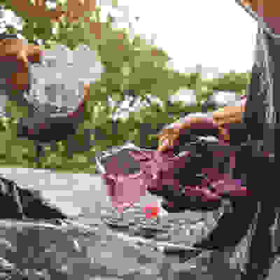 A person holds a crumpled clear plastic bottle in one hand and a purple plastic bottle in the other, surrounded by discarded materials.