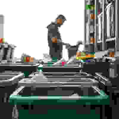 Various recycling bins are arranged in a room, with a person sorting through items in the background. Bright natural light fills the space.
