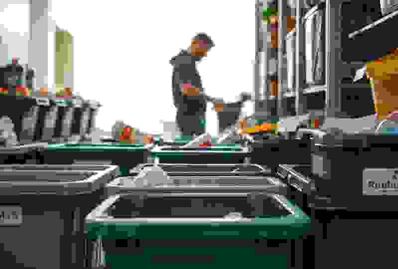 Various recycling bins are arranged in a room, with a person sorting through items in the background. Bright natural light fills the space.