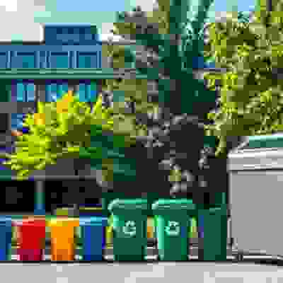 A row of colorful recycling bins and a gray waste container are positioned in a green park area, with trees and a modern building in the background.