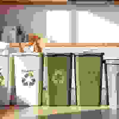 A bright kitchen backdrop features a four-bin system: recycling bins labeled for paper, plastic, and metal, alongside a gray trash can.