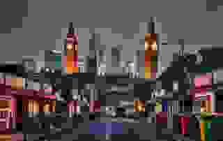 A twilight view of Big Ben surrounded by modern skyscrapers, with a bustling street lined with shops and colorful trash bins.